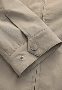 Manga de chaqueta color beige claro con un botón y cierre a presión. Presenta una textura suave, costuras visibles y un borde ajustado.