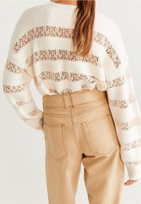 Cremefarbener Strickpullover mit offenen Spitzeneinsätzen, kombiniert mit hoch taillierten beigen Hosen mit Gesäßtaschen und geradem Bein.