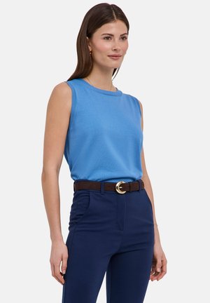 Vrouw draagt een lichtblauw mouwloos topje, ingestopt in marineblauwe broek met een donkerbruine riem en gouden gesp, staand tegen een effen achtergrond.