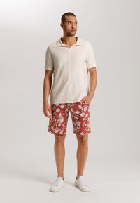 Beige Ripp-Polohemd mit kurzen Ärmeln, kombiniert mit rot gemusterten Shorts. Getragen mit weißen Sneakers und vor hellem Hintergrund stehend.
