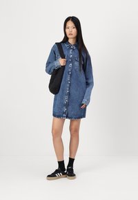 Vestito camicia in denim blu medio, maniche lunghe, davanti con bottoni, due tasche a petto, indossato con una borsa nera e scarpe sportive nere.