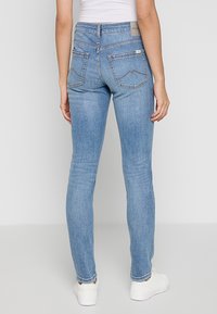 Jean en denim bleu ajusté avec une texture lisse, comportant deux poches arrière avec un design de couture courbée et une petite étiquette de marque au niveau de la taille.