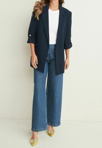 Mujer vistiendo un blazer azul marino con mangas arremangadas, top blanco, jeans de piernas anchas azules y tacones amarillos de punta con hebillas de joyas.