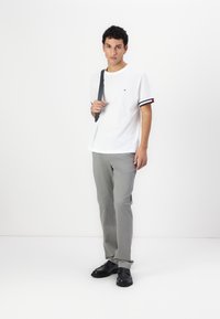 Hvid bomulds t-shirt med sorte og røde striber ved ærmerne, parret med grå bukser. Modellen har sorte sko på og holder en grå rem.