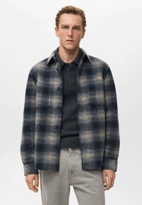 Kockovaná flanelová košeľa v tmavo modrej a béžovej farbe, s gombíkovým zapínaním, šírokým golierom a náprsné vrecko. Nosená cez tmavú pletenú polokošeľu.