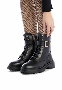 Botas de tobillo de cuero negro con cordones y cremallera, herrajes dorados, acentos de tachuelas y suela gruesa. Terminadas con un cuello acolchado.