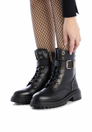 Botas de tobillo de cuero negro con cordones y cremallera, herrajes dorados, acentos de tachuelas y suela gruesa. Terminadas con un cuello acolchado.