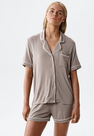 Młoda kobieta ubrana w taupe krótkie piżamy zapinane na guziki z białym obszyciem, stojąca na tle jednolitego, jasnego tła.