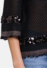 Schwarzes Strickoberteil mit kurzen Ärmeln, das ein strukturiertes offenes Gewebe und Paillettenakzente an den Bündchen und dem Saum aufweist.