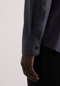 Manica di camicia button-up grigia con tessuto testurizzato, caratterizzata da un bottone scuro e dettagli a contrasto nel polsino. Mano posizionata vicino all'orlo.