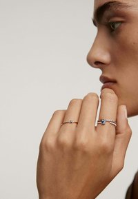 Dos anillos en una mano: un anillo de oro con una piedra azul y otro anillo de plata con piedras preciosas azules y transparentes, sobre un fondo neutro.