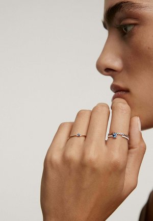 Dos anillos en una mano: un anillo de oro con una piedra azul y otro anillo de plata con piedras preciosas azules y transparentes, sobre un fondo neutro.