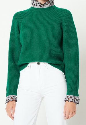 Femme portant un pull côtelé vert par-dessus un chemisier blanc à pois avec des poignets festonnés, associé à un pantalon blanc taille haute.
