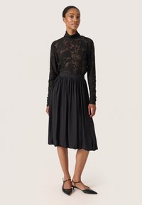 Top in pizzo nero a maniche lunghe con motivo floreale, abbinato a una gonna midi plissettata nera. Il modello indossa mocassini neri a punta.