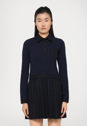 Marineblå langermet polo med krage, med en svart lomme; kombinert med en svart plissert skjørt med tynne vertikale striper.