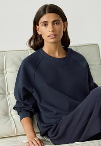 Junge Frau mit glattem dunklem Haar, die goldene Creolen und einen dunkelblauen Sweatshirt trägt, sitzt auf einem weißen gepolsterten Ledersofa.