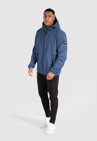 Veste coupe-vent bleue avec capuche, fermeture éclair à l'avant, poches latérales et manches élastiques ; associée à un pantalon noir et des chaussures blanches.