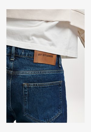 Jeans in denim di colore blu scuro con una finitura testurizzata. Presentano un'etichetta in pelle marrone con la scritta "2Y STUDIOS" impressa sulla vita posteriore.