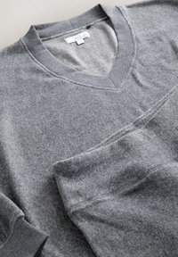 Pelēks V veida džemperis, izgatavots no mīkstas, teksturētas auduma. Tam ir ribota apkakle un piedurkņu maliņas, gluda apdare un saskaņoti šuvumu detaļas.