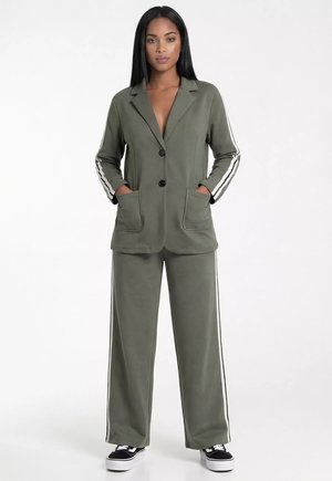Kobieta stoi z rękami w kieszeniach, ubrana w oliwkowo zieloną marynarkę na dwa guziki oraz pasujące szerokie spodnie z białymi paskami po bokach i czarne trampki.