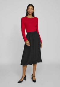 Röd, ribbad långärmad tröja med guldaxelackcenter, matchad med en svart midikjol med midjeband och svarta klackskor. Släta texturer.