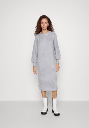 Robe en maille grise avec des manches longues et bouffantes et un col rond. Le tissu présente une texture côtelée. Associée à des bottines blanches à la cheville.