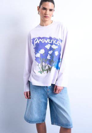 Γυναίκα φοράει λευκό μακρυμάνικο μπλουζάκι με το κείμενο "Provence" και λευκά λουλούδια σε μπλε φόντο, συνδυασμένο με ανοιχτόχρωμα τζιν σορτς.