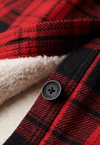 Rødt og sort ternet stof med sort knap mærket "Superdry" og synligt hvidt fleeceforetunder materialet.