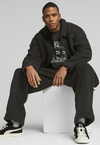 Svart fleece-jacka och byxset med en texturerad yta. Under finns en grafisk svart t-shirt med ett cirkulärt mönster. Svarta sneakers med vita accenter.
