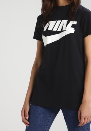 Kobieta w czarnym krótkim t-shircie z dużym białym logo Nike i niebieskich dżinsach, stojąca na jednolitym jasnym tle.