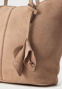 Μπεζ τσάντα tote από σουέτ με δύο λαβές, που διαθέτει δεμένη λεπτομέρεια και αντίθετη ραφή. Λεία υφή και δομημένος σχεδιασμός.