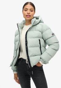 Chaqueta de plumas verde claro con capucha, que presenta un zipper negro, bolsillos laterales y puños de canalé. Llevada sobre un suéter crema y jeans negros.