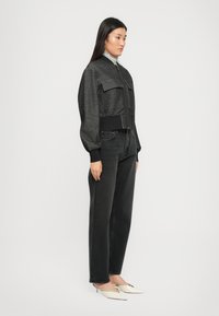 Veste noire coupée avec deux poches avant, ourlet côtelé et manches larges, associée à un jean foncé et des chaussures pointues de couleur crème.