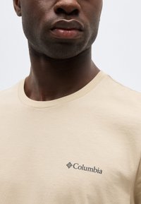 Κοντινό πλάνο προσώπου που φορά μια μπεζ μπλούζα Columbia με στρογγυλή λαιμόκοψη και το λογότυπο ορατό στο στήθος, πάνω σε ένα απλό φόντο.