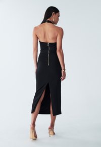 Crna halter midi haljina s oblikovanim siluetom, zlatni detalj zatvarača na leđima i bočni prorezi koji se protežu do sredine teladi.