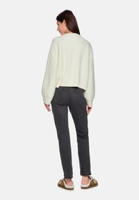 Gehäkelter cremefarbener Pullover mit einem cropped Design, kombiniert mit schmalen, anthrazitgrauen Jeans und braunen Slippern aus Kunstfell.