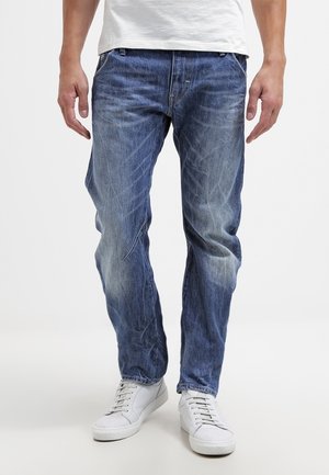 Homme portant un jean bleu délavé et des baskets blanches, debout, les bras relâchés le long du corps sur un fond blanc uni.