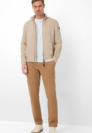 Mann trägt beige Reißverschlussjacke, weißes T-Shirt, braune Hose und weiße Sneakers, steht mit einer Hand in der Tasche vor weißem Hintergrund.