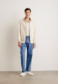 Beige bomber dzseki bordázott mandzsettával, fehér pólóval viselve, kék farmernadrággal és fehér sportcipővel párosítva; sima textúra.