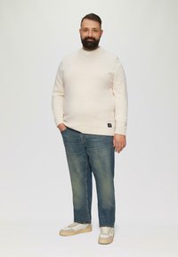 Helle beige strukturierte Pullover, blaue Jeans und hellfarbene Sneaker. Das Model steht mit den Händen in den Taschen vor einem einfarbigen Hintergrund.
