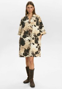 Korte mouw jurk in beige met een bruine en zwarte koeienprint, met een kraag en een losse pasvorm, gecombineerd met bruine kniehoge laarzen.