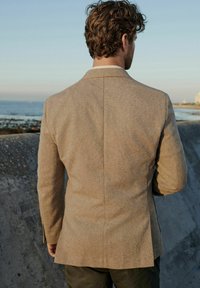 Blazer marrón con textura y corte ajustado, con una abertura trasera sencilla y solapas con muesca, visto desde la parte de atrás contra un fondo costero.