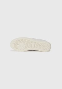 Hvit sneakersåle med teksturert gummi og mønstrede spor i beige, med en sirkulær logo i midten og side detaljer i rødt.