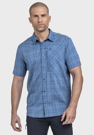 Mann trägt ein kurzärmliges, blaues Hemd mit dezentem Karomuster und Brusttasche, steht vor einem schlichten Hintergrund.