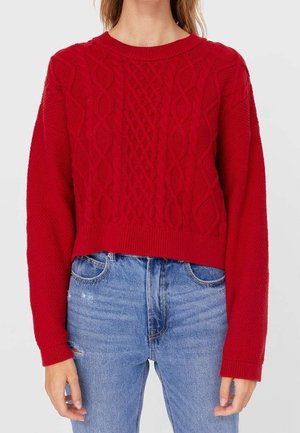 Woman wearing a red cable-knit sweater with long sleeves and light blue high-waisted jeans with slight distressing on left thigh.