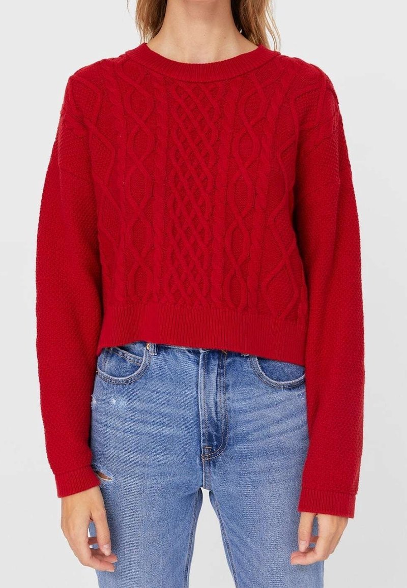 Femme portant un pull rouge en maille torsadée à manches longues et un jean bleu clair taille haute légèrement usé sur la cuisse gauche.