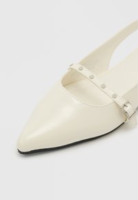 Άσπρο μυτερό επίπεδο παπούτσι slingback με ρυθμιζόμενο λουράκι με αγκράφα και ασημένια τρουκς στην κορυφή, μαύρη σόλα ορατή στην εμπρόσθια άκρη.