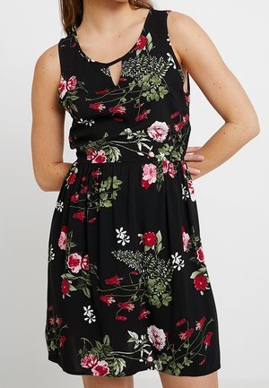 Mouwloze zwarte jurk met roze, rode en witte bloemmotieven, nauwsluitende taille en klein keyhole-halslijn, gedragen door een persoon met de hand op de heup.