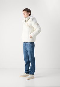 Hettejakke i hvitt med en stor logo foran, med hette og sidelommer. Kombinert med blå jeans og hvite joggesko.