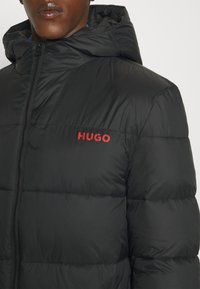 Schwarze, wattierte Jacke mit hohem Kragen, vorne mit Reißverschluss und rotem "HUGO"-Logo auf der Brust. Glatte Textur mit gestepptem Design.